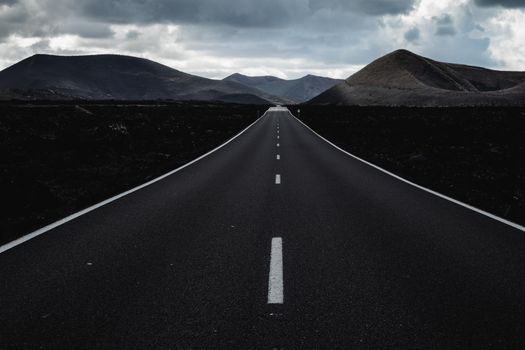 Endless road on a volcano in Timanfaya National Park in Lanzarote in the Canary Islands with a continuous line, black volcanic rocks on the side and volcanoes in mist in background