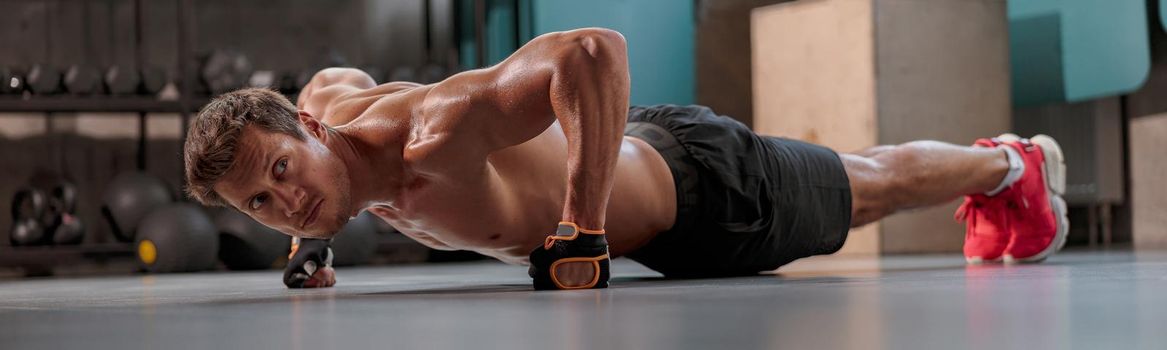 Strong muscular man in training gloves and shorts performing sports exercise push-up from the floor in the fitness club