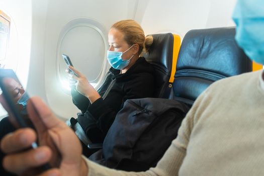 A young couple wearing face mask, using smart phone while traveling on airplane. New normal travel after covid-19 pandemic concept