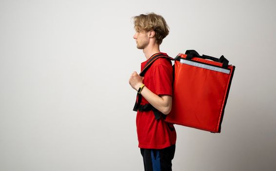 Young courier in a red uniform t-shirt and with red food thermo bag on a shoulder standing isolated on white background studio. Food delivery service