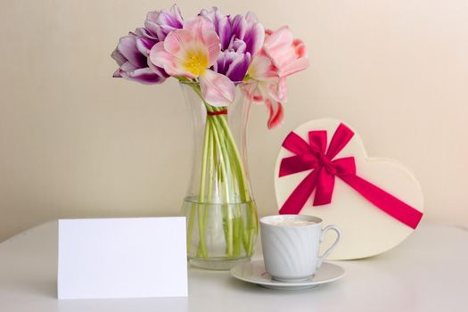 A bouquet of open pink and purple tulips, a gift box in the shape of a heart, with a pink bow, a white cup and a blank card with space for text on a white table in a bright room.
