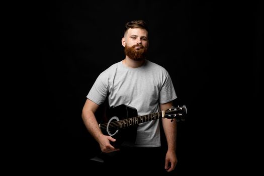 Handsome brunette bearded man musician standing and holding a acoustic guitar in a hand and looks in a camera on a black background studio. Ready to play a music