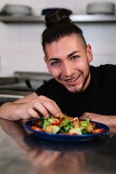 portrait of a young man chief, long hair tied back, dressed in a black polo shirt and a dark striped apron, putting ingredients on a salad plate in a professional kitchen, on a table full of food. The atmosphere is cold and clean, with the backdrop of professional aluminium tables and the fires of the restaurant kitchen. looking at camera