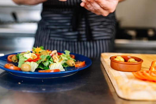 Detail of the hands of a young man, dressed in a black polo shirt and a dark striped apron, placing a salad on a blue plate in a professional kitchen, on a table full of food. The atmosphere is cold and clean, with the backdrop of professional aluminium tables and the fires of the restaurant kitchen.