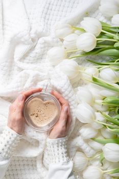 hands of two women holding a heart-shaped cup with cappuccino on two sides, female friendship, flat lay with white tulips, mother's day, valentine's day,