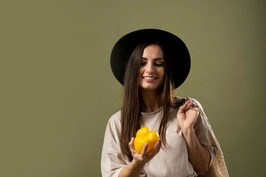 Eco friendly smiling woman in beige t-shirt and black hat holding reusable cotton eco bags full of groceries on a shoulder and yellow pepper