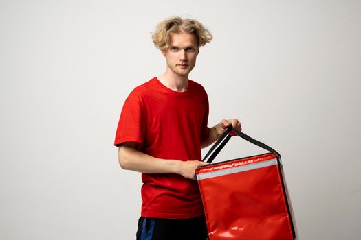 Young courier opening thermal bag on white background. Food delivery service. Delivery guy in a red t-shirt uniform work as courier and holds red thermal food backpack. Service concept