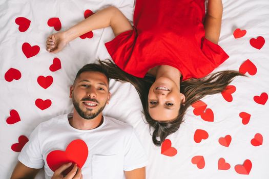 Top view of happy young couple in love with red paper hearts lying on the white bed. Love and relationship. High quality photo