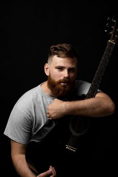 Portraite of handsome brunette bearded man musician, guitarist holding a acoustic guitar in a hand and looks in a camera on a black background studio. Ready to play a music