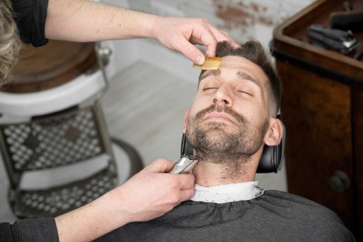 Young hipster Caucasian man during beard grooming in modern barber shop. Men's hair styling. Handsome man getting new hairstyle with electric trimmer. High quality photography