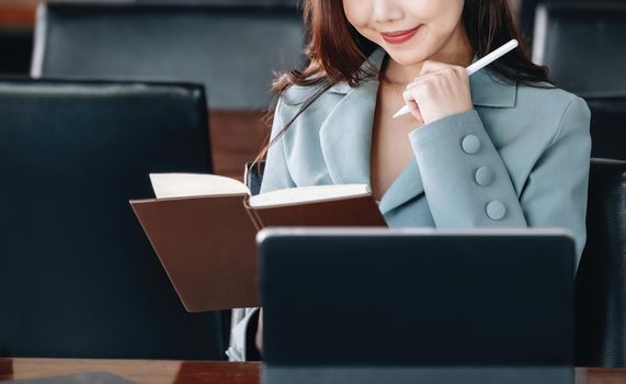 Asia woman entrepreneur or businesswoman showing a smiling face while reading a book developing financial and investing strategies and operating a computer tablet working on a wooden table