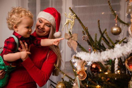 Mom is carrying her little son in her arms and he is pointing his finger at the Christmas tree with a bow on it. A woman wearing a red Santa hat.