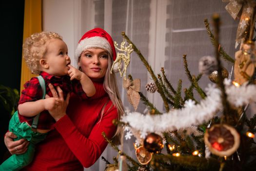A woman with her son in her arms stands by a decorated tree for Christmas. Woman wearing a red Santa hat.
