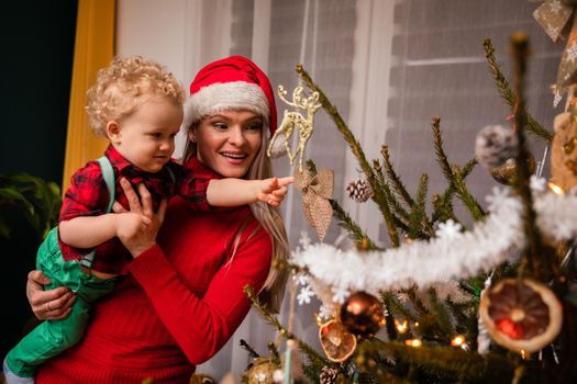 Mom is carrying her little son in her arms and he is pointing his finger at the Christmas tree with a bow on it. A woman wearing a red Santa hat.