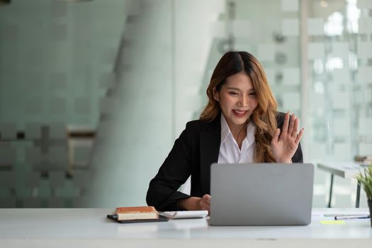 Portrait of smiling asian woman waving hello talking on video call. Successful young woman sitting white suits. Business conference via laptop.