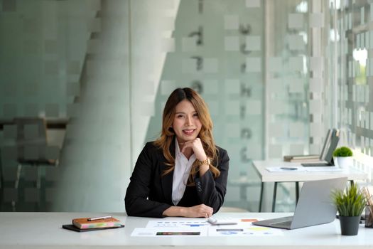 Portrait of attractive business woman sitting at her desk with laptop computer in office. Smiling business woman in black suit at office.