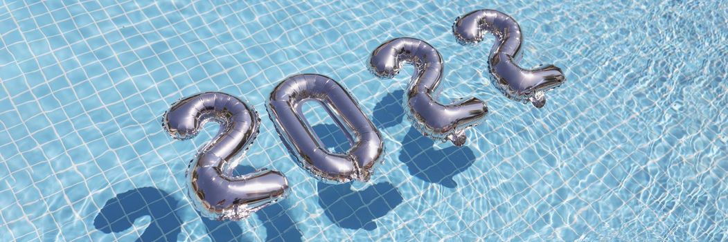 Top view of silver numbers 2022 float across blue water of swimming pool. Preparing decoration for home party in pool, festive holiday. New year, new period, celebration concept