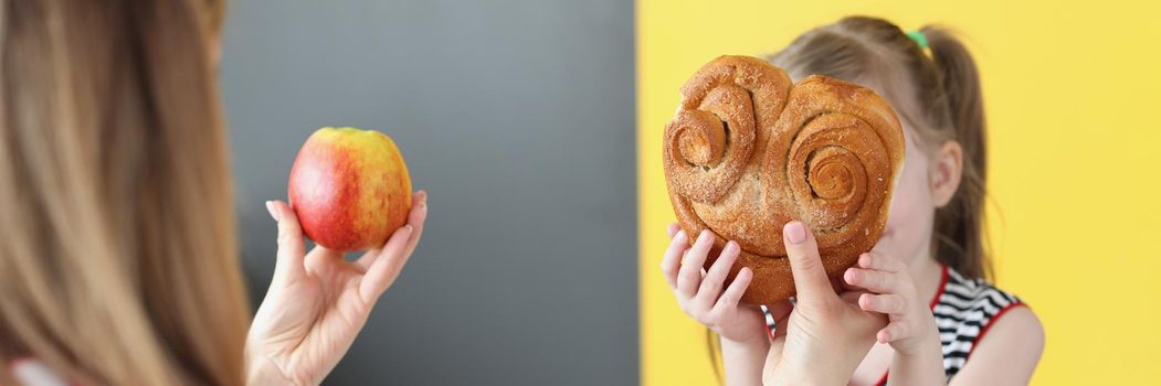 Close-up of little girl playing with mum, kid holding big bun, mum want to change bun for apple. Mother serve proper food for child. Healthy eating, diet, childhood, family time, fun concept