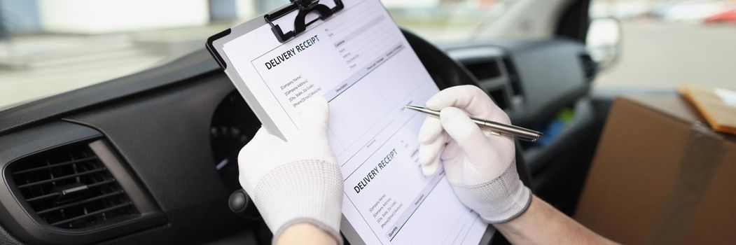 Close-up of courier driver in medical protective gloves holds documents with delivery receipt, fill in paper with pen in car, contract of cooperation. Delivery service concept