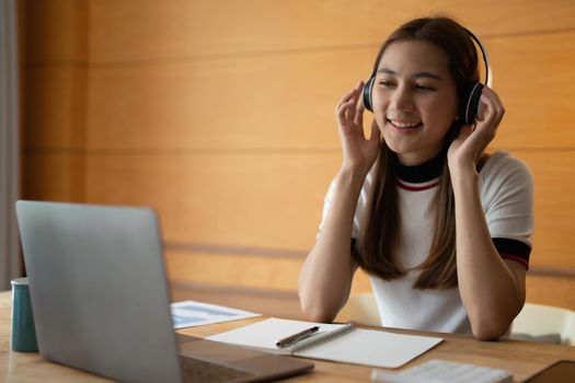 Smiling asian girl student wear wireless headphone study online, happy young woman learn language listen lecture watch webinar write notes look at laptop at home, distant education.