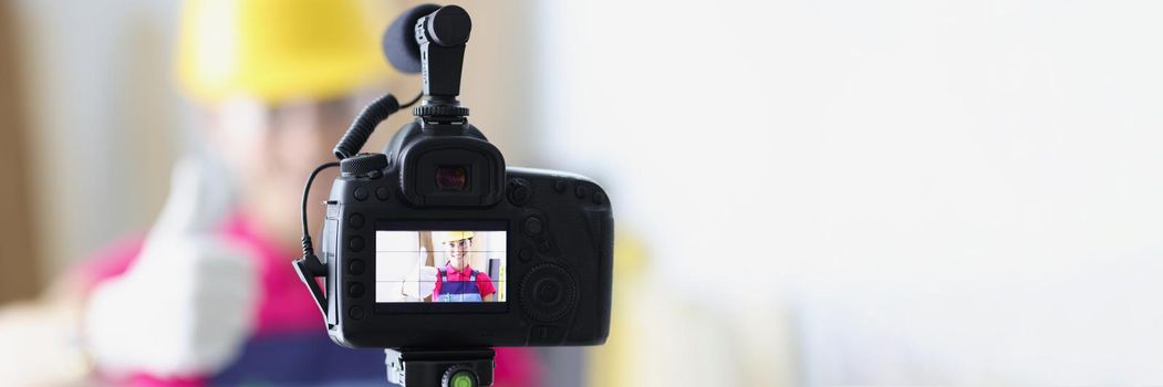 Close-up of videocamera with female repairer in helmet showing thumb up gesture to camera. Building blogging and vlogging concept. Redecoration online idea