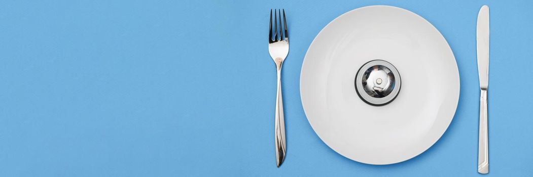 Top view of white plate with iron bell and appliances for food. Time to eat, dieting and meal-planning concept. Meals on schedule idea. Isolated on blue background