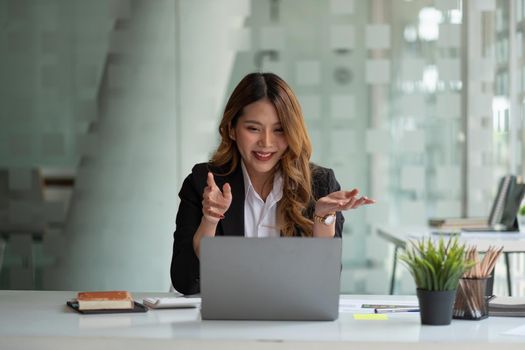 Portrait of smiling asian woman waving hello talking on video call. Successful young woman sitting white suits. Business conference via laptop.