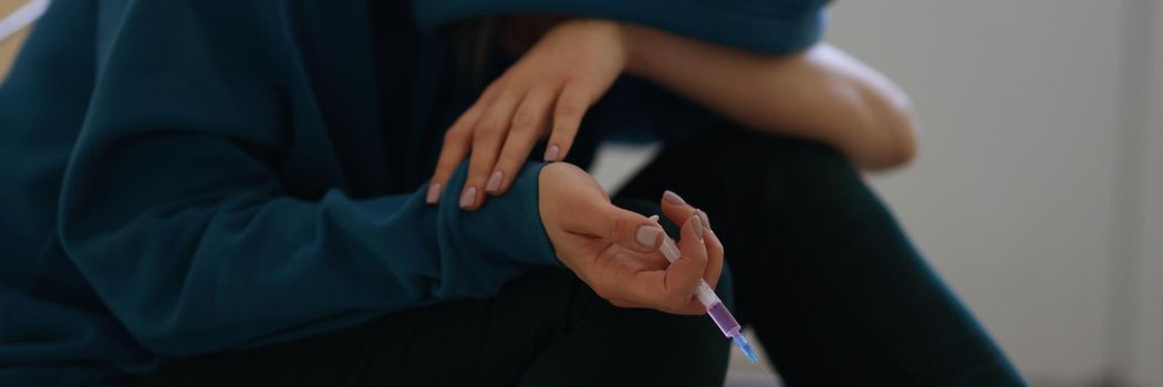 Close-up of female in hoodie holding syringe and getting high. Drug addict person stoned after narcotic injection sitting on floor. Drug habit and meth-head concept