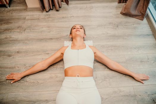 Adult athletic woman, in white bodysuit, performing fascia exercises on the floor - caucasian woman using a massage foam roller - a tool to relieve tension in the back and relieve muscle pain - the concept of physiotherapy and stretching training.