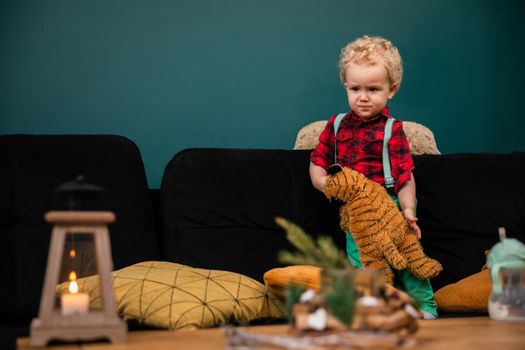 A small child stands with a scowling face on a bed with a toy in his hand. A little boy with blue eyes and curly blond hair.