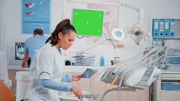 Stomatologist watching monitor with horizontal green screen while assistant bringing teeth radiography for examination. Dentist looking at x ray and display with chroma key for mockup template