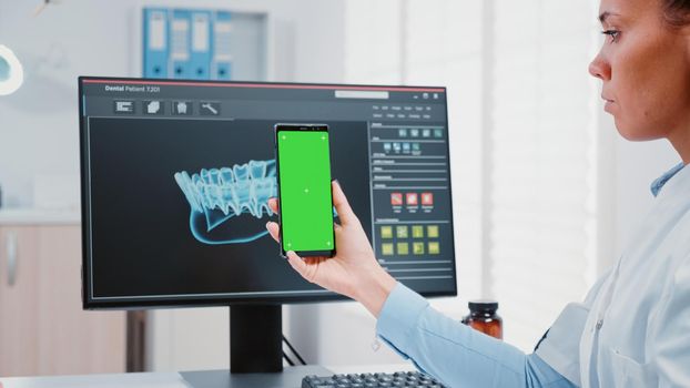 Stomatologist vertically holding green screen on mobile phone in dentist office. Dental specialist looking at modern device with isolated background and mockup template for teethcare