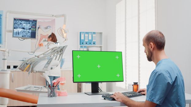 Man working with keyboard and computer with green screen on desk in dentistry office. Oral care assistant using mockup template and isolated background in dental cabinet for teethcare