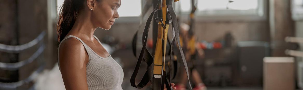 Side view of young female wearing workout clothes exercising with suspension trainer on blurred background of gym