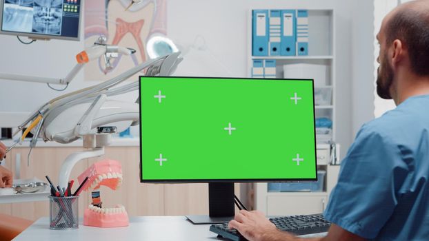 Man nurse looking at horizontal green screen on monitor in dental office while dentist working with dentistry equipment. Assistant using computer with chroma key and isolated template