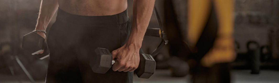 Cropped view of muscular young man holding dumbbells in hands and doing exercise to strengthen his biceps in fitness center