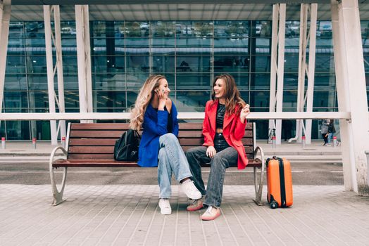 Two happy girls sitting on a bench near airport, with luggage. Air travel, summer holiday.