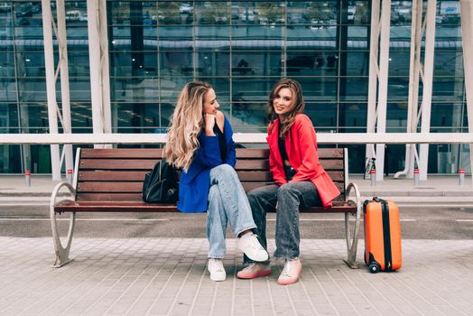 Two happy girls sitting on a bench near airport, with luggage. Air travel, summer holiday.