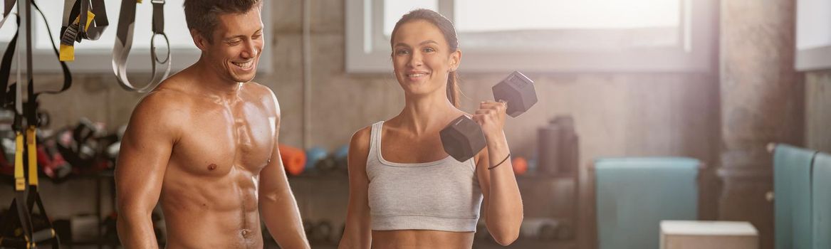 Slender pretty lady holding dumbbells in her hands performing sports exercise in the gym with smiling strong man