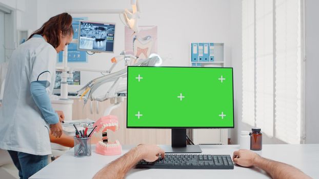 POV of man using keyboard and computer with green screen at desk in oral care cabinet. Assistant looking at monitor with isolated background and mockup template for teethcare and dental checkup