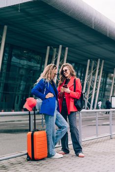 Two happy girls using smartphone checking flight or online check-in at airport together, with luggage. Air travel, summer holiday, or mobile phone application technology concept