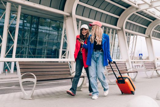 Two happy girls using smartphone checking flight or online check-in at airport together, with luggage. Air travel, summer holiday, or mobile phone application technology concept