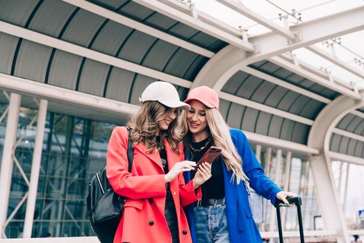 Two happy girls using smartphone checking flight or online check-in at airport together, with luggage. Air travel, summer holiday, or mobile phone application technology concept