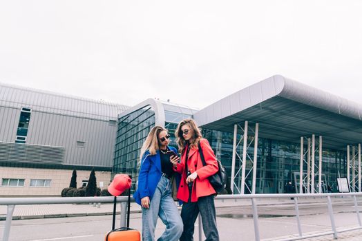 Two happy girls using smartphone checking flight or online check-in at airport together, with luggage. Air travel, summer holiday, or mobile phone application technology concept