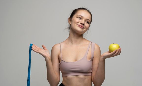 Skeptical sexy brunette women in sport outfit standing with a measuring tape and holding a apple on a white background. Beauty and health concept. Healthy eating and diet