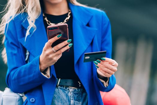 Happy fashion blonde shopper wearing a blue jacket paying on line with a credit card and a smart phone on the street.