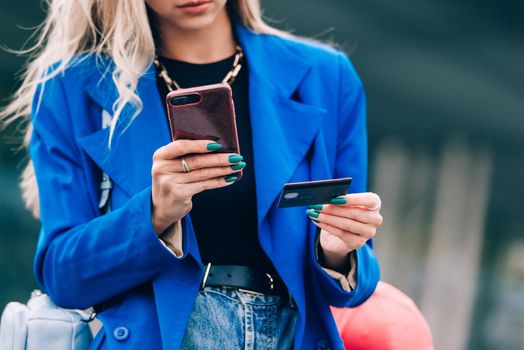 Happy fashion blonde shopper wearing a blue jacket paying on line with a credit card and a smart phone on the street.