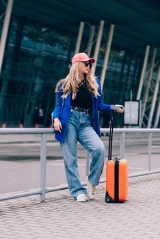 Portrait of a traveler student standing with an orange suitcase near an airport. Young fashionable woman in a blue jeans and jacket, black shirt and white sneakers. Pink baseball cap