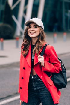 glamour woman in trendy outfit posing against the building urban background, fashion look. Outdoor fashion portrait of stylish young woman wearing black jeans, red jacket, top and a cap