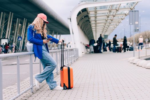 Portrait of a traveler student standing with an orange suitcase near an airport. Young fashionable woman in a blue jeans and jacket, black shirt and white sneakers. Pink baseball cap